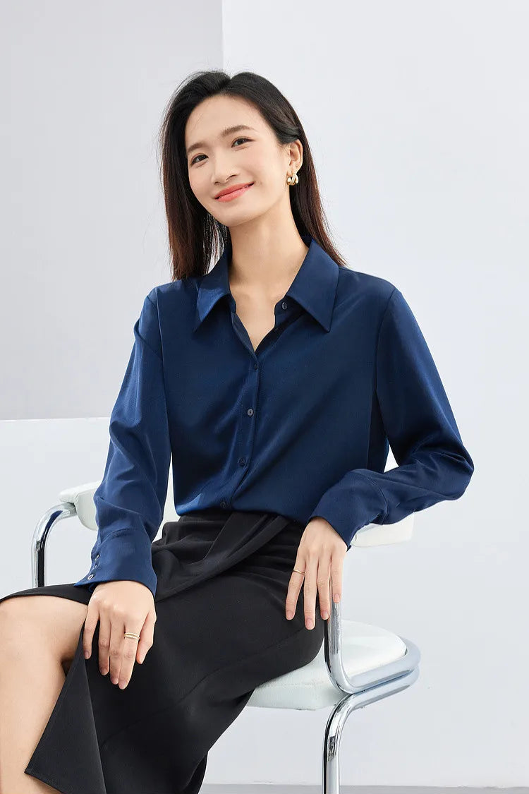 Woman wearing a blue shirt and black skirt sitting on a white chair against a light gray background.